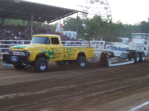 1994 Ford F-150 - A vintage yellow Ford pickup truck, customized with flame graphics, competes in a pull event, dragging a weighted sled.