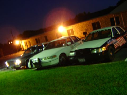 1988 Ford LTD Crown Victoria - Three police cars are parked in a line at night, with one an American flag decal on its fender.