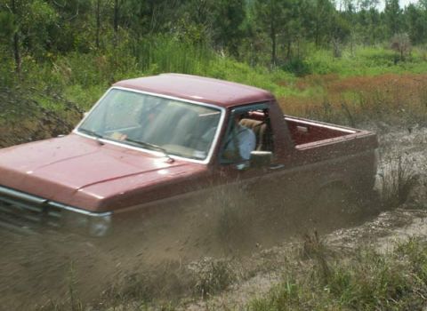 1988 Ford Bronco - A red pickup truck navigates through a muddy terrain, kicking up dirt and water.