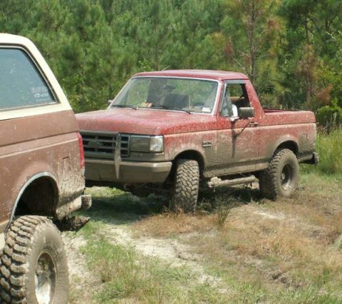 1988 Ford Bronco - Two off-road vehicles are parked on a dirt path surrounded by trees.