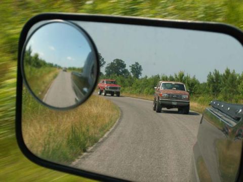 1988 Ford Bronco - Two trucks drive down a rural road, one brown and one red, as seen in a side mirror.