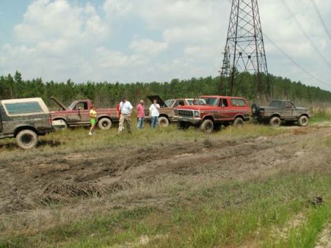 1988 Ford Bronco - A group of off-road vehicles are gathered in a muddy field on a cloudy day, with a large power line tower visible.