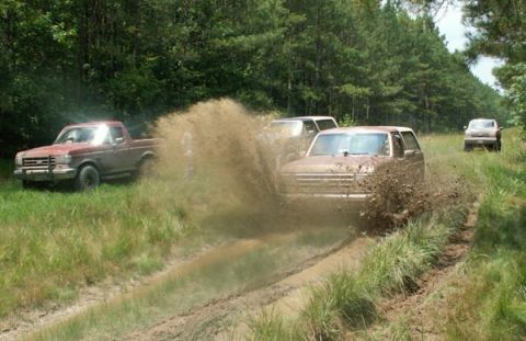 1988 Ford Bronco - Several SUVs are navigating a muddy trail, kicking up large plumes of dirt and water.