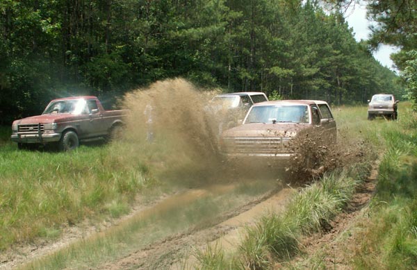 Mud-splattered 1987 Ford Bronco driving through a muddy track with trees in the background