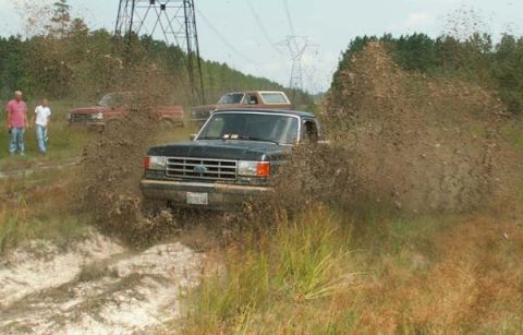 1988 Ford Bronco - Multiple trucks navigate a muddy track, kicking up large plumes of dirt.
