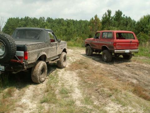 1988 Ford Bronco - Two Ford Broncos, one muddy and one red, drive off-road on a dirt track with trees in the background.
