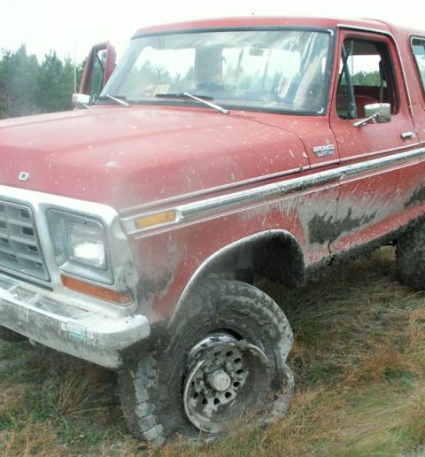 1988 Ford Bronco - A red Ford Bronco is stuck in mud with a damaged front wheel, its doors open.