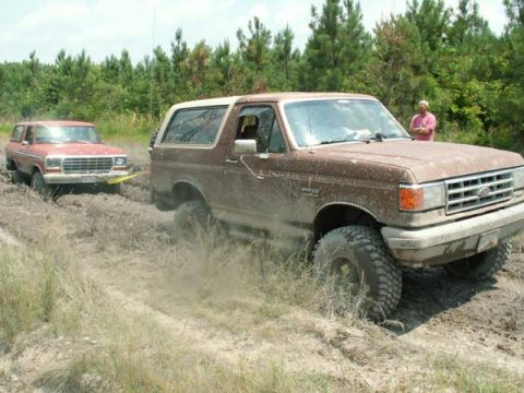 1988 Ford Bronco - Two Ford Broncos are stuck in the mud on a trail with trees in the background.