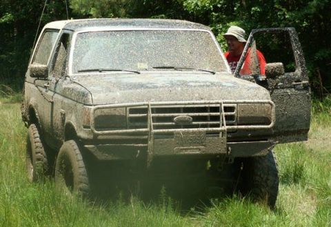 1988 Ford Bronco - This heavily mud-caked SUV is parked in a grassy, wooded area with its driver's side door ajar.