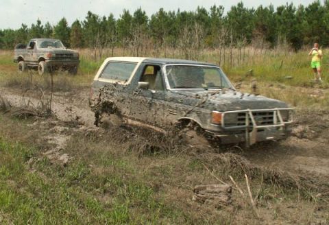 1988 Ford Bronco - Two trucks navigate a challenging muddy terrain, kicking up significant spray as they go.