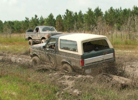 1988 Ford Bronco - Two Ford Broncos navigating a muddy off-road course with trees in the background.