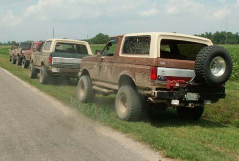 1988 Ford Bronco - Several Ford Broncos are lined up on a dirt road, kicking up dust as they drive past a grassy field under a cloudy sky.