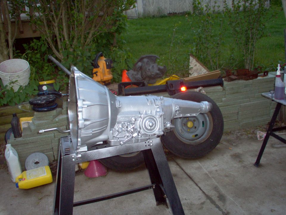 Silver-painted Ford automatic transmission on a black stand outdoors, with a wheel and yard items behind it