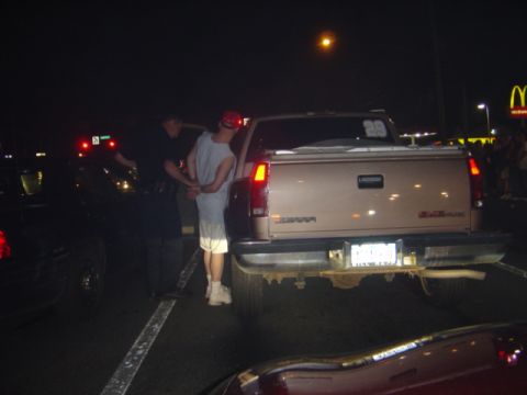 1998 Ford Taurus - A man is handcuffed by a police officer next to a GMC pickup truck in a parking lot at night.