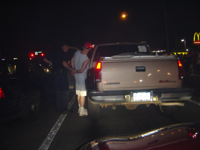 Rear view of a tan GMC pickup truck with a man in handcuffs beside it at night