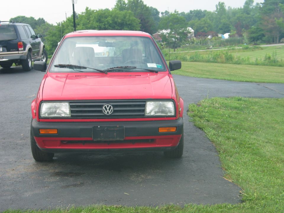 Red Volkswagen Golf parked on asphalt driveway, front view. Another SUV is visible to the left