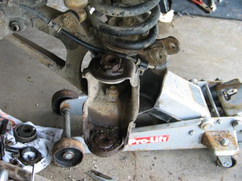1995 Ford Bronco - Front suspension torn down on a concrete garage floor, showing a coil spring, radius arm and greasy cast axle knuckle bracket supported by…
