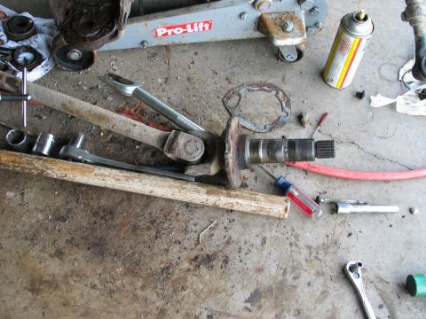 1995 Ford Bronco - Overhead shot of a driveline and suspension teardown laid out on a cracked concrete garage floor: a rusty splined axle shaft with its flang…