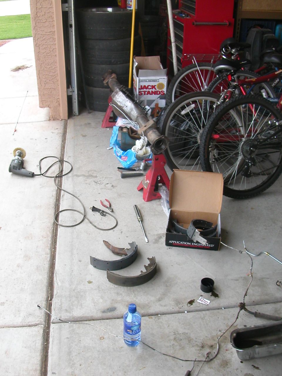 Tools and parts laid out on garage floor, including brake shoes, a grinder, and a catalytic converter on jack stands