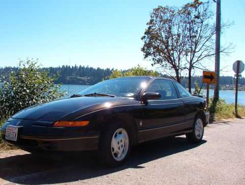 1981 Ford Bronco - Saturn SC coupe parked roadside with a lake view. The album claims 1981 Ford Bronco.