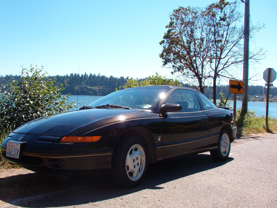 Black Saturn SC coupe with white alloy wheels parked beside a waterway