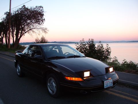 1981 Ford Bronco - A black coupe with its pop-up headlights illuminated sits by the water during a colorful sunset.