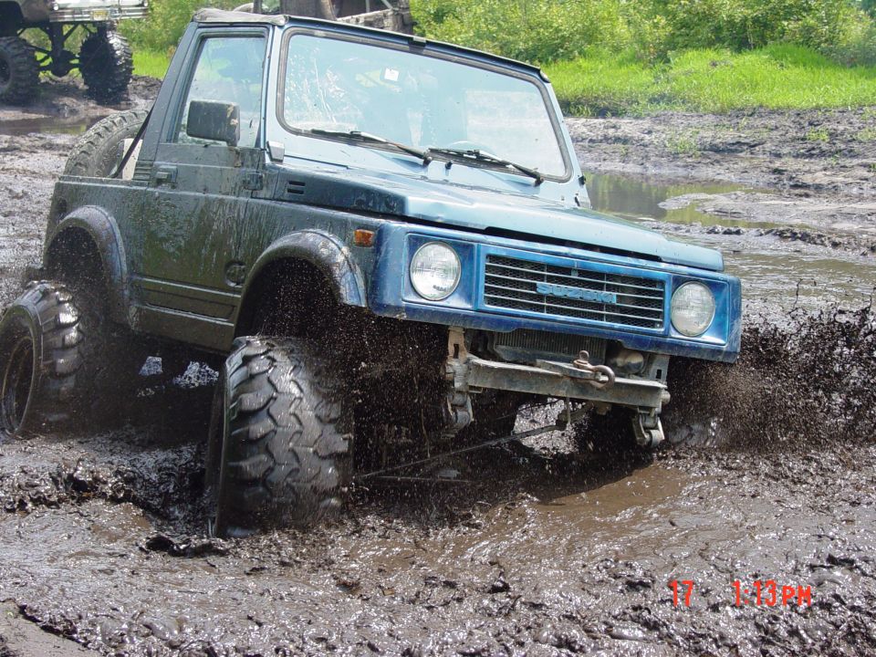 Blue Suzuki Samurai off-roading in mud, front passenger side tire deep in muck. Another vehicle is visible in the background