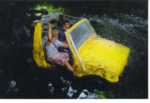 1987 Suzuki Samurai - Two people are in a yellow off-road vehicle partially submerged in dark, murky water.