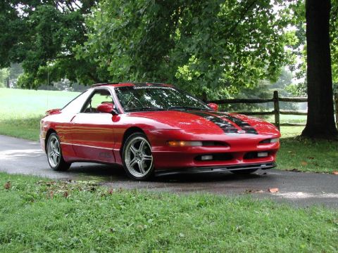 1997 Ford Probe - A red 1997 Ford Probe with black racing stripes is parked on a wet road, surrounded by trees and a wooden fence.
