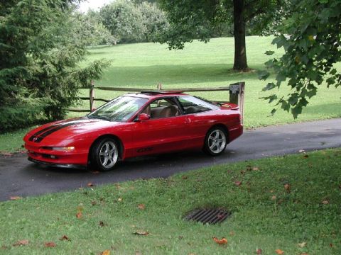 1997 Ford Probe - A red sports car with racing stripes is parked on a driveway with a grassy lawn and trees in the background.