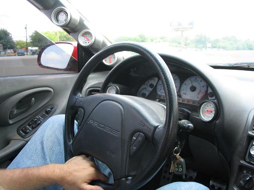Driver's perspective inside a car, showing steering wheel with 'PROBE' text, dashboard gauges, and a side mirror
