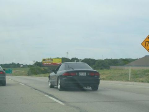 1994 Ford Probe - A black 1994 Ford Probe is seen from behind while driving on a highway under an overcast sky.