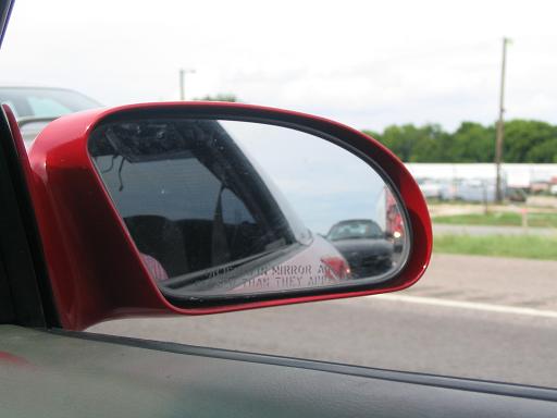 Red car side mirror reflecting a black car on a road
