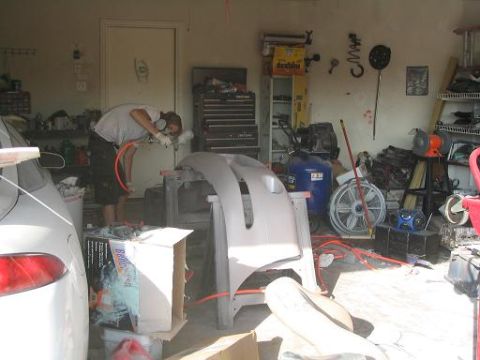 1994 Ford Probe - A person spray paints a car bumper in a cluttered garage with tools and equipment visible.
