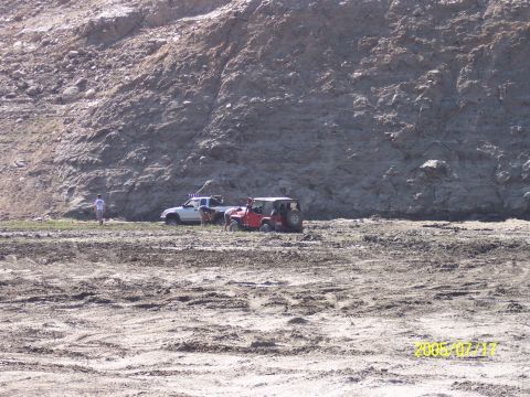 1985 Ford Bronco - Two off-road vehicles are parked on muddy terrain with a large rock face in the background.
