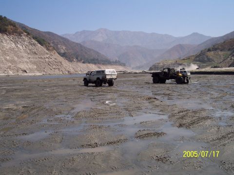 1985 Ford Bronco - Two off-road vehicles are parked in a dry riverbed with mountains in the background.