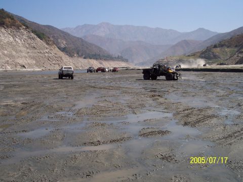 1985 Ford Bronco - Several off-road vehicles traverse a muddy, dry riverbed with mountains in the distance under a clear blue sky.