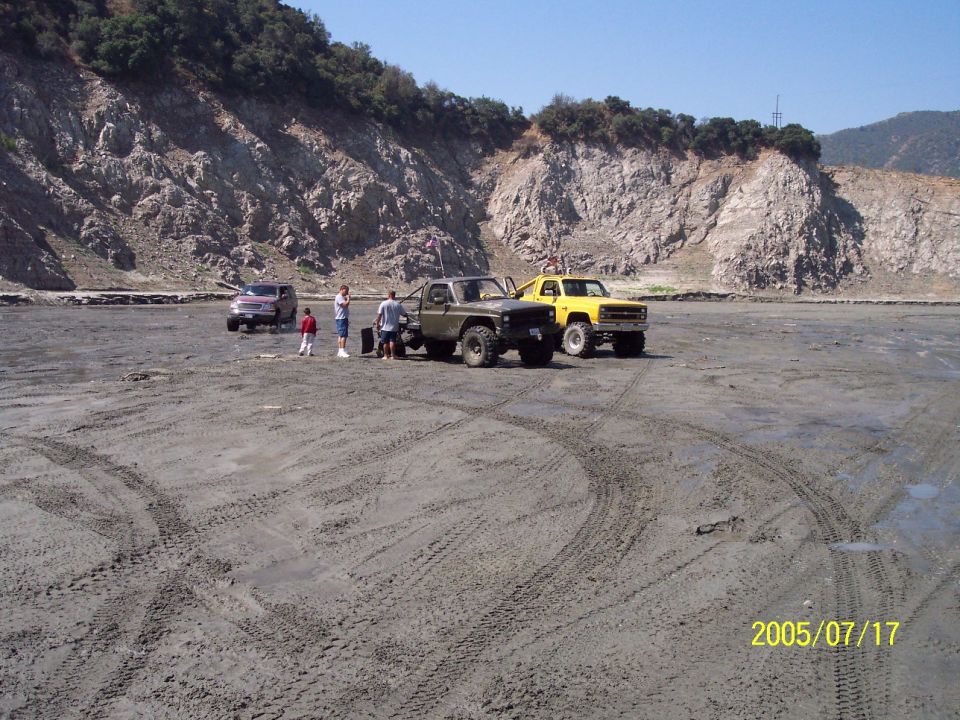 Three off-road vehicles and two people stand in a dry, dusty area with tire tracks; rocky hills in background