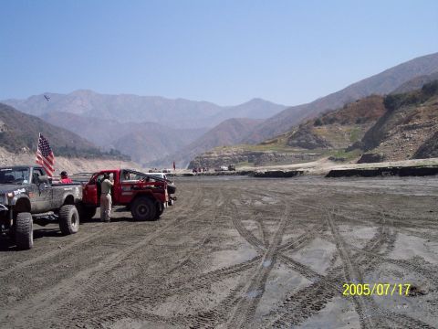 1985 Ford Bronco - Off-road vehicles are parked in a dry riverbed with mountains in the distance under a clear blue sky.