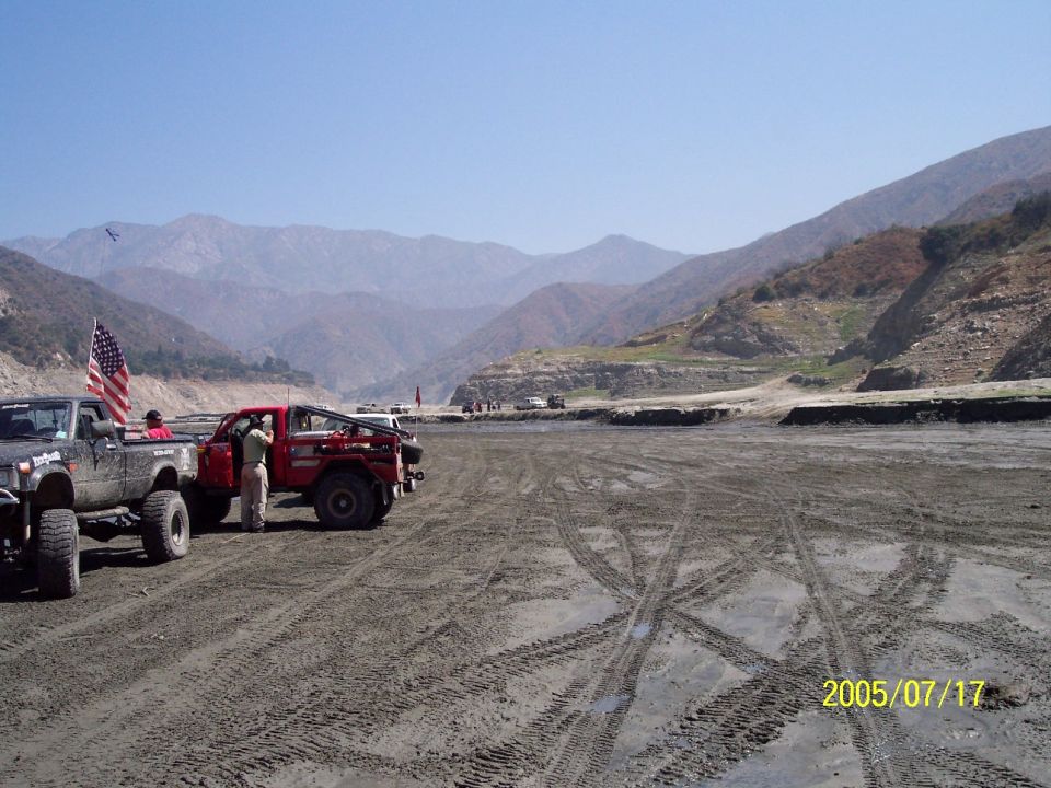 Two off-road trucks, one red and one dark, parked on a dirt surface with tire tracks, mountains in background