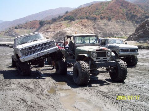 1985 Ford Bronco - Multiple off-road vehicles navigate a muddy, uneven landscape under a bright sky.
