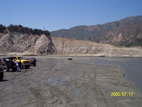 1985 Ford Bronco - Multiple off-road vehicles are parked and driving in a wide, muddy area with tire tracks visible.