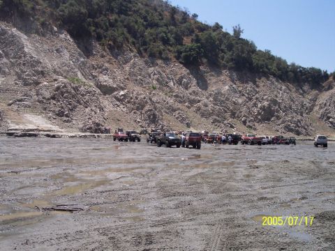 1985 Ford Bronco - A group of off-road vehicles are gathered on muddy terrain with a large rock face in the background.