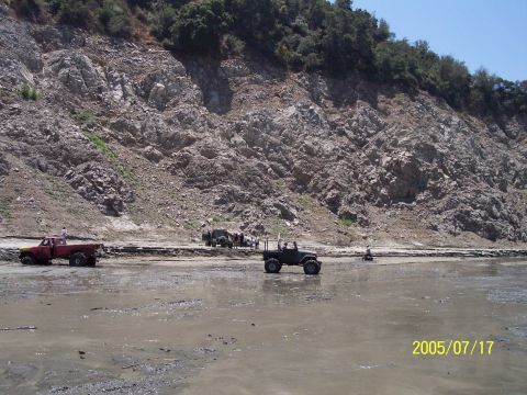 1985 Ford Bronco - Several off-road vehicles are parked and driving in a muddy, rocky terrain with a steep, vegetated cliff face in the background.