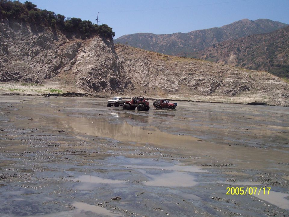Three lifted 4x4 trucks parked in a shallow muddy wash below dry chaparral hills