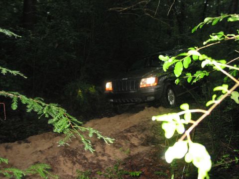 2000 Jeep Grand Cherokee - A dark 2000 Jeep Grand Cherokee navigates a muddy path through a dense forest at dusk.