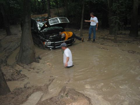2000 Jeep Grand Cherokee - A black Dodge Ram truck is stuck in deep mud with its hood open as people attempt to fix it.