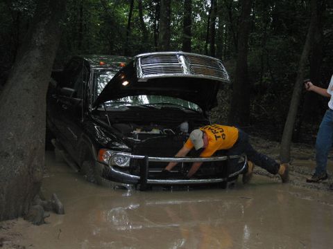 2000 Jeep Grand Cherokee - Man works on a black Dodge Ram stuck in mud with hood open, while another person watches.