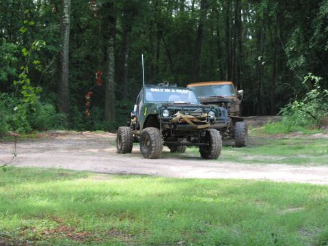 2000 Jeep Grand Cherokee - A heavily modified Jeep with oversized tires and a humorous windshield decal sits parked in a wooded area next to a truck.