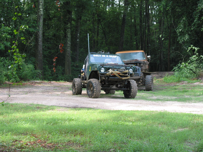 Modified green Jeep with large tires and "ONLY IN A HEAP" on windshield, parked next to a truck